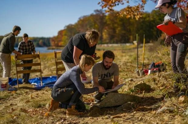 People at work on archaeological dig site, trees and bay in background