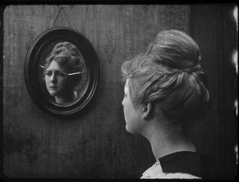Black and white still photo shows woman with hair up, looking sadly into a cracked mirror.
