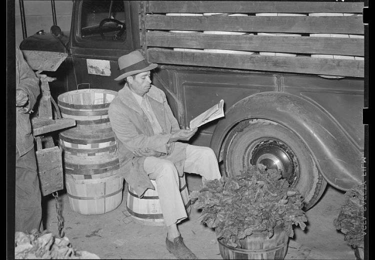 Man reading a newspaper next to a truck and baskets of spinach