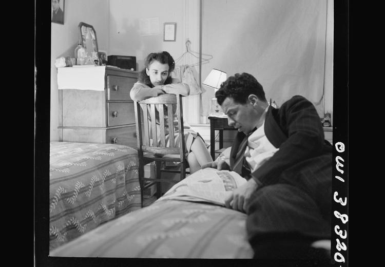 Man reading newspaper aloud in foreground. Woman listening in background.